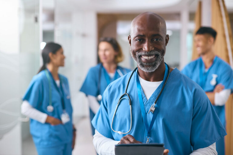 Portrait Of Mature Male Doctor With Digital Tablet In Hospital With Colleagues In Background