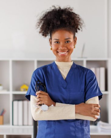 Medical professional in blue scrubs working on laptop, woman smiling while standing in well-lit healthcare environment